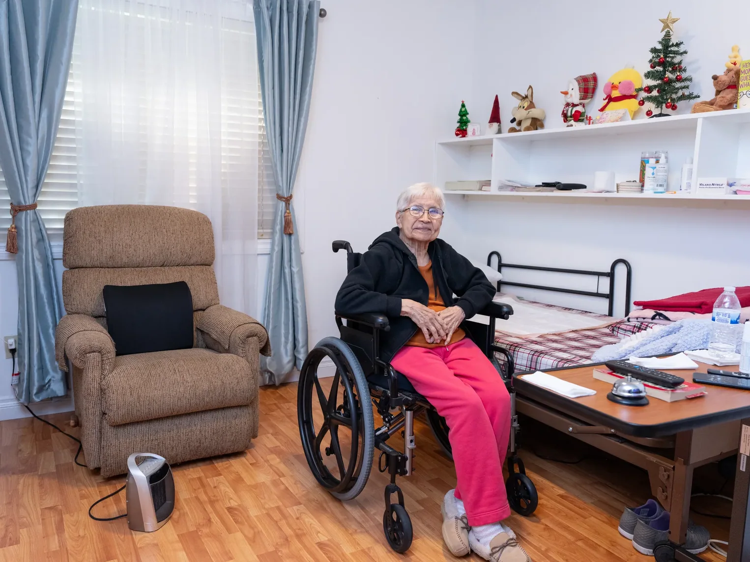 Senior resident seated in wheelchair in well-lit private bedroom with hospital bed and personal decorations